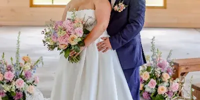 A bride and groom standing together in a rustic wooden venue with floral arrangements and white drapery.