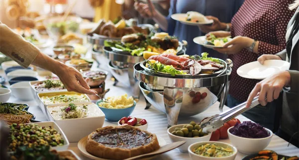 A catering buffet table filled with various food dishes in chafing dishes and bowls, with people serving themselves.