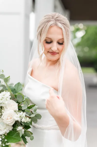 A bride with styled blonde hair and makeup holding a bouquet of white flowers and greenery.