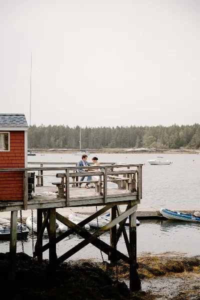A bride and groom standing on a wooden dock by the water with a red boathouse and boats in the background.