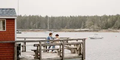 A bride and groom standing on a wooden dock by the water with a red boathouse and boats in the background.