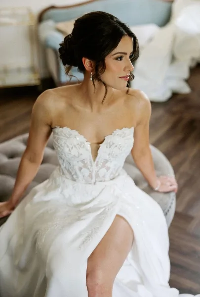 A bride wearing a white lace wedding dress with a styled updo and professional makeup, seated in a room with a blue sofa in the background.