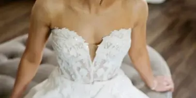 A bride wearing a white lace wedding dress with a styled updo and professional makeup, seated in a room with a blue sofa in the background.