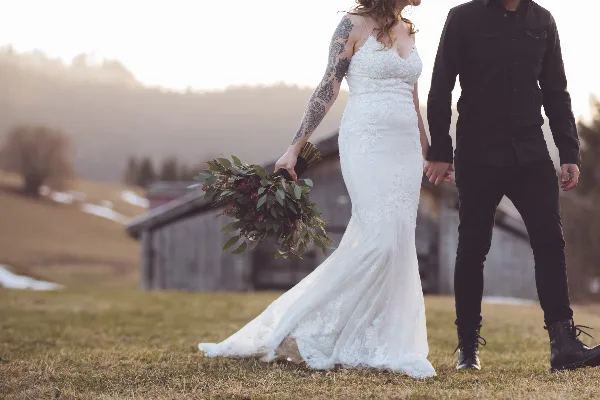 A bride in a white lace gown and a groom in a black shirt walking hand-in-hand in a grassy field with a rustic barn in the background.