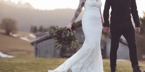 A bride in a white lace gown and a groom in a black shirt walking hand-in-hand in a grassy field with a rustic barn in the background.
