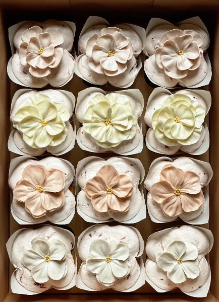 A box of twelve cupcakes decorated with large, piped buttercream flowers in white and pale pink.