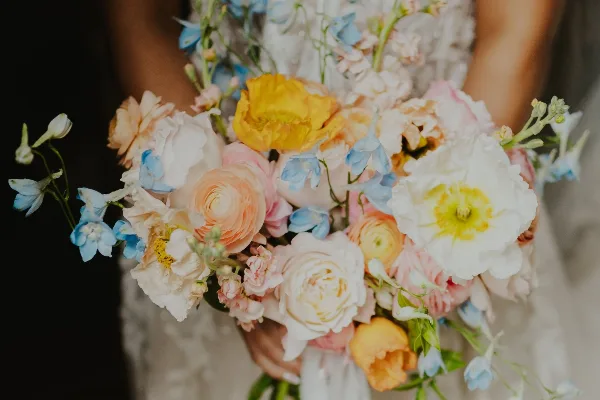 A close-up photograph of a wedding bouquet featuring peach ranunculus, white roses, and blue delphiniums.