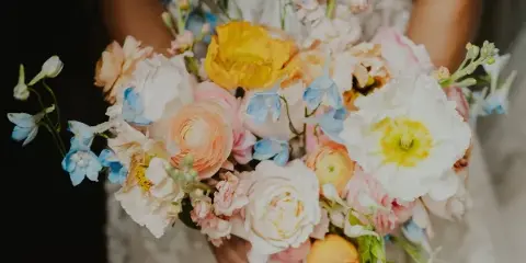 A close-up photograph of a wedding bouquet featuring peach ranunculus, white roses, and blue delphiniums.