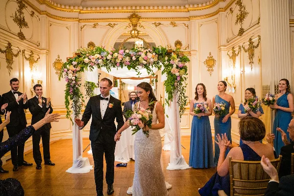 A wedding couple walking down the aisle past a large floral arch and bridesmaids holding bouquets.