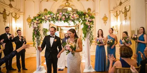 A wedding couple walking down the aisle past a large floral arch and bridesmaids holding bouquets.