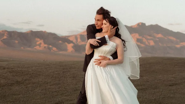 A bride and groom embracing in a field with mountains in the background.