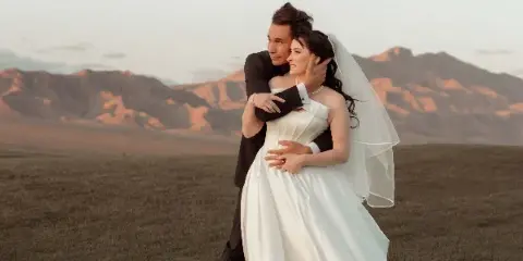A bride and groom embracing in a field with mountains in the background.