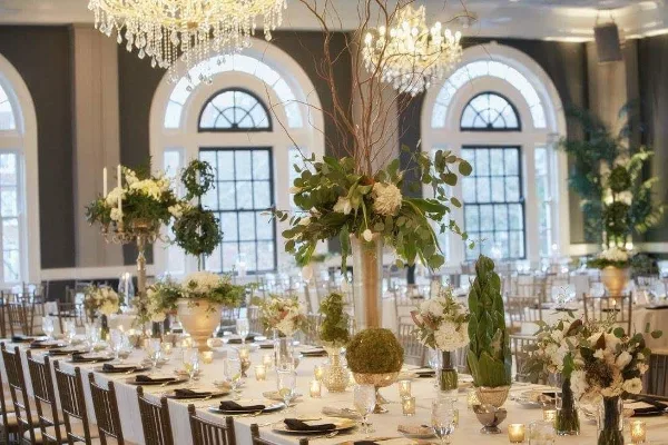 A formal wedding reception table setting featuring tall floral centerpieces, greenery, and candlelight in a grand ballroom.