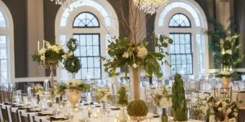 A formal wedding reception table setting featuring tall floral centerpieces, greenery, and candlelight in a grand ballroom.