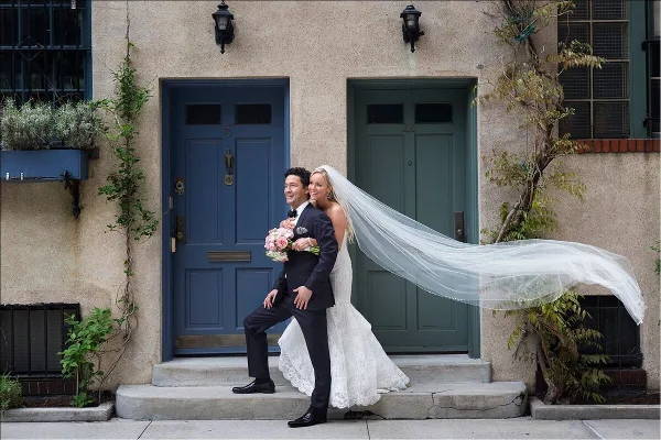 A bride and groom posing on the steps of a New York City brownstone, with the bride's long veil blowing in the wind.