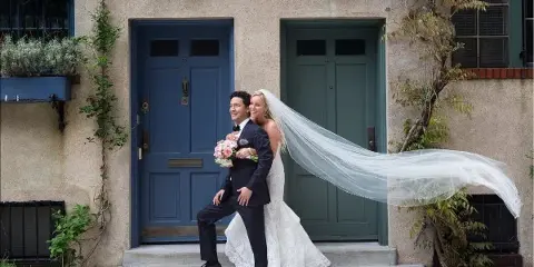 A bride and groom posing on the steps of a New York City brownstone, with the bride's long veil blowing in the wind.
