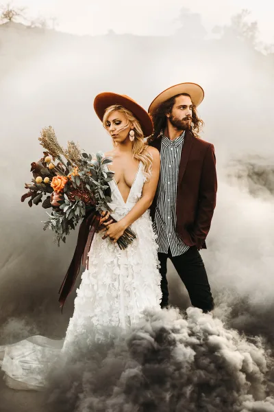 A bride and groom posing in a studio setting with smoke effects, the bride wearing a white floral dress and holding a bouquet.