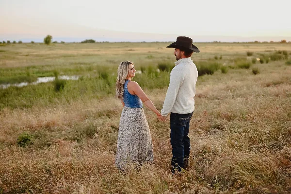 A couple holding hands in a grassy field at sunset, with the man wearing a cowboy hat.