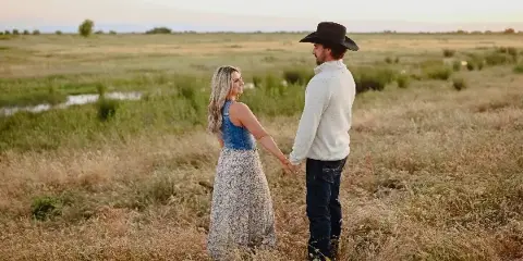 A couple holding hands in a grassy field at sunset, with the man wearing a cowboy hat.