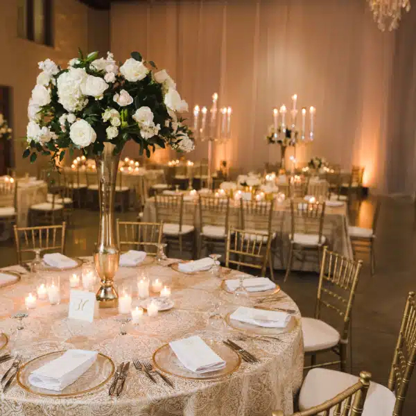 A formal event space featuring round tables with white tablecloths, gold chiavari chairs, and large floral centerpieces.