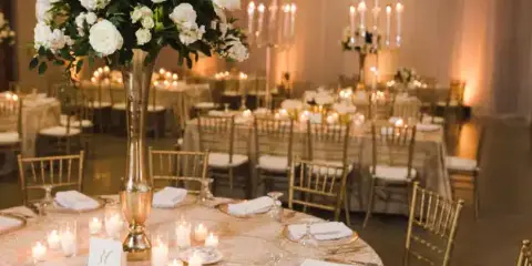 A formal event space featuring round tables with white tablecloths, gold chiavari chairs, and large floral centerpieces.