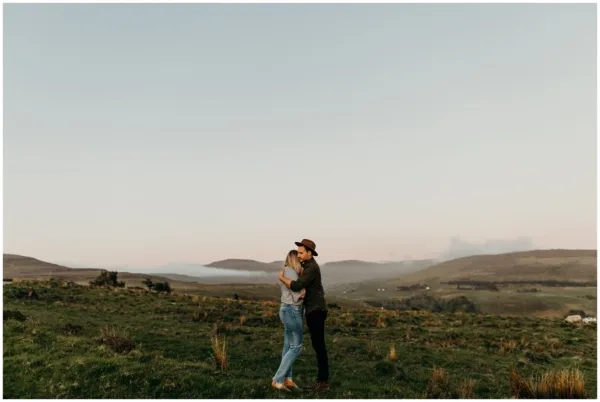 A couple embracing in a grassy field with rolling hills and a soft sky in the background.