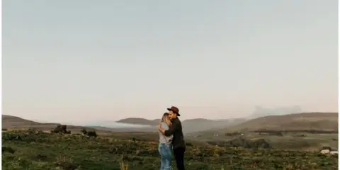 A couple embracing in a grassy field with rolling hills and a soft sky in the background.