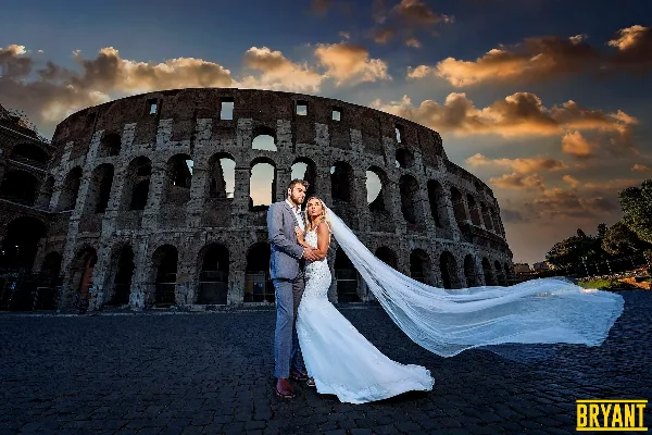 A bride and groom posing in front of the Colosseum in Rome, Italy, with the bride's veil blowing in the wind.