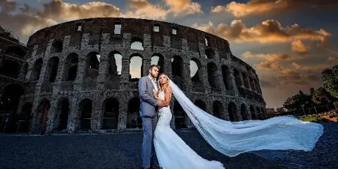 A bride and groom posing in front of the Colosseum in Rome, Italy, with the bride's veil blowing in the wind.
