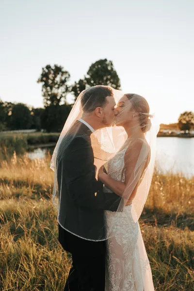 A bride and groom sharing a kiss in a grassy field with a lake and trees in the background.