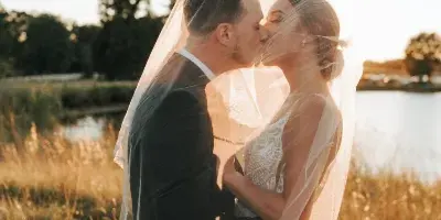 A bride and groom sharing a kiss in a grassy field with a lake and trees in the background.