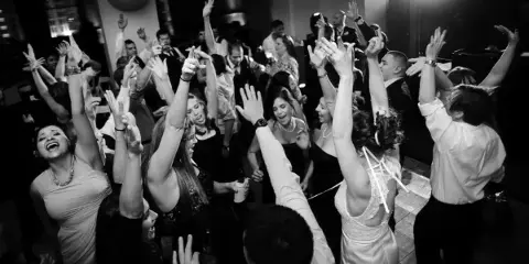 A black and white photo of a crowded wedding dance floor with guests raising their hands and dancing.