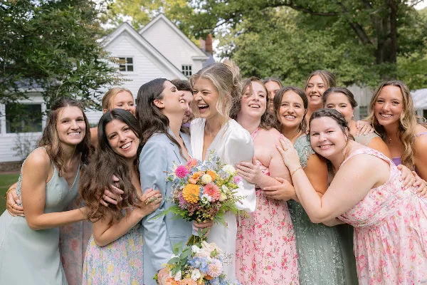 A bride and her bridesmaids smiling and laughing together outdoors at a wedding.