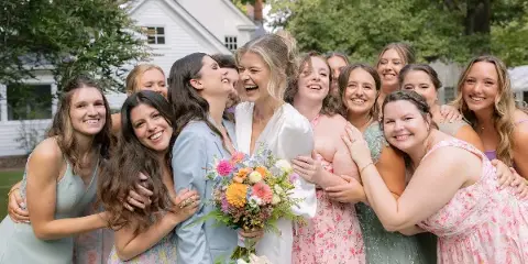 A bride and her bridesmaids smiling and laughing together outdoors at a wedding.