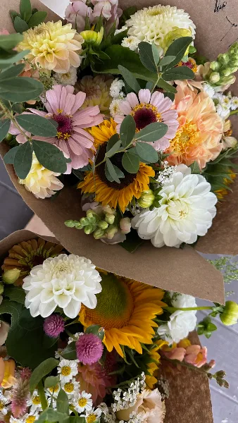 A large, colorful bouquet of mixed cut flowers including daisies, sunflowers, and dahlias, wrapped in brown paper.