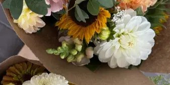 A large, colorful bouquet of mixed cut flowers including daisies, sunflowers, and dahlias, wrapped in brown paper.