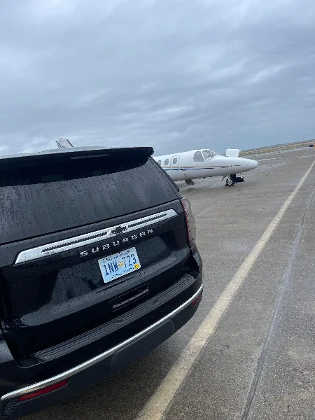 Rear view of a black Chevrolet Suburban with a Rhode Island Public Service license plate parked on an airport tarmac near a small aircraft.