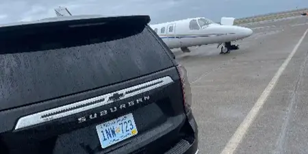 Rear view of a black Chevrolet Suburban with a Rhode Island Public Service license plate parked on an airport tarmac near a small aircraft.