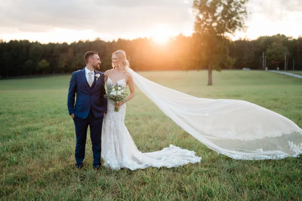 A bride and groom standing in a grassy field at sunset, with the bride's long veil blowing in the wind.