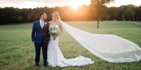 A bride and groom standing in a grassy field at sunset, with the bride's long veil blowing in the wind.