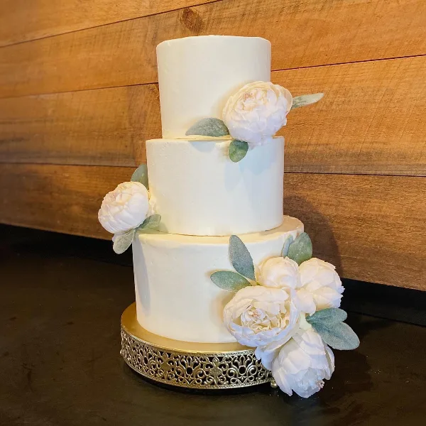 Three-tier white frosted wedding cake with white flowers and eucalyptus leaves on a gold cake stand against a wooden wall.