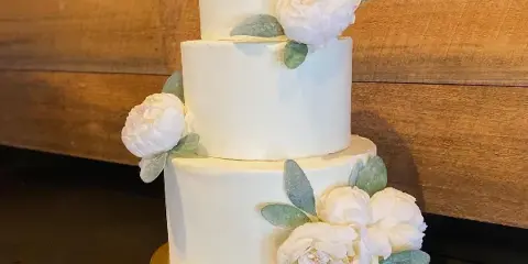 Three-tier white frosted wedding cake with white flowers and eucalyptus leaves on a gold cake stand against a wooden wall.