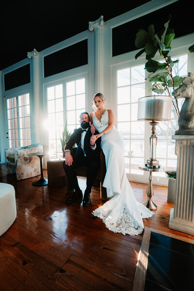 A bride and groom posing in a moody, editorial style wedding photograph inside a venue with large windows.