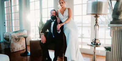 A bride and groom posing in a moody, editorial style wedding photograph inside a venue with large windows.