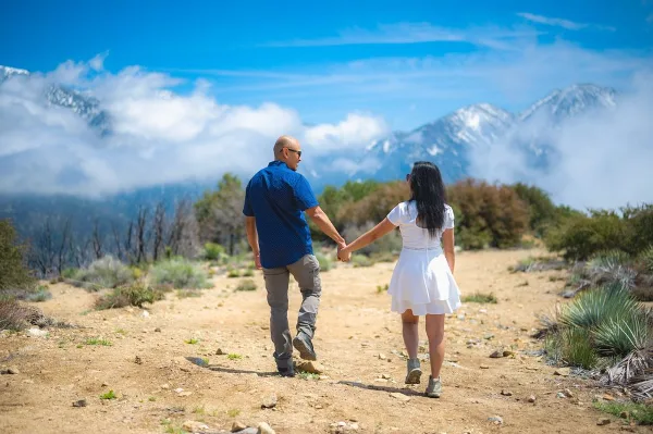 A couple holding hands walking on a dirt path with a mountain landscape in the background.