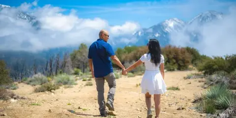 A couple holding hands walking on a dirt path with a mountain landscape in the background.