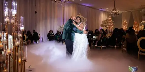 A bride and groom dancing at a wedding reception in a ballroom with chandeliers and floor fog.