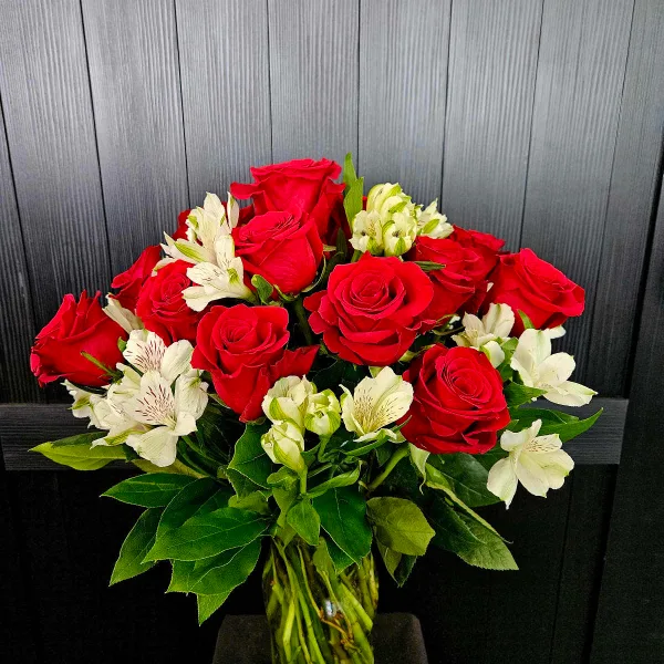 A bouquet of red roses and white alstroemeria in a clear glass vase against a dark wood background.