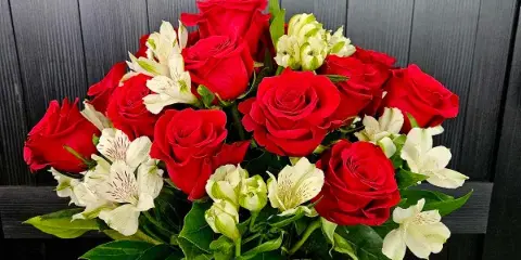 A bouquet of red roses and white alstroemeria in a clear glass vase against a dark wood background.