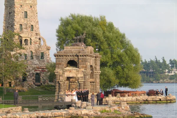 A wedding party poses in front of the stone Dove-Cote structure with the main tower of Boldt Castle visible in the background.